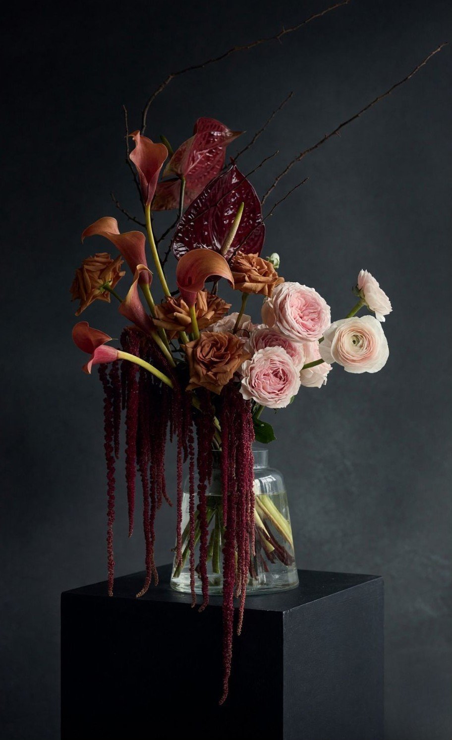 Elegant floral arrangement in glass vase with deep red calla lilies, burgundy roses, and pale pink garden roses on black pedestal against dark background