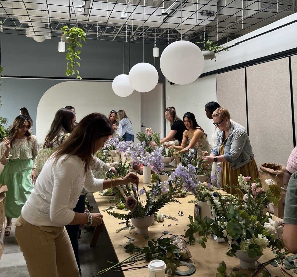 Group of women attending a floral arrangement workshop in a bright studio with hanging spheres and potted flowers on a wooden table
