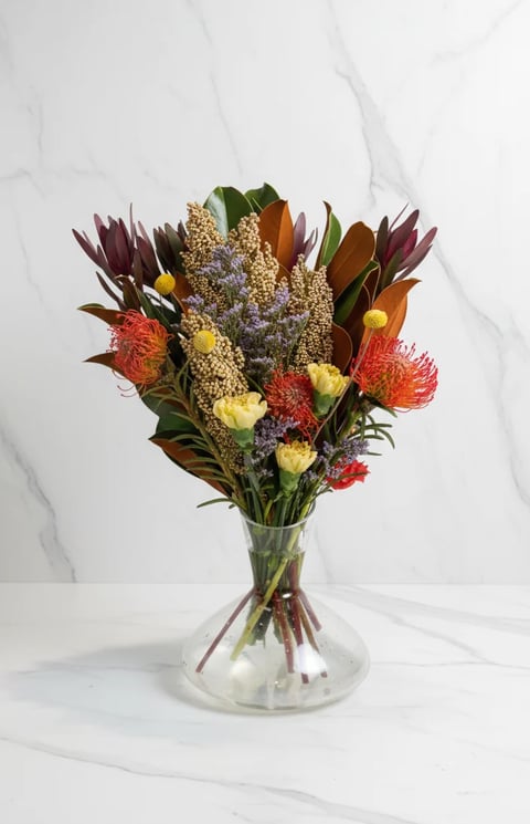 Colorful flower bouquet in glass vase with red proteas, yellow blooms, and burgundy foliage on white marble background