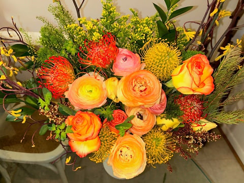 Vibrant floral arrangement with orange and coral ranunculus, red protea, yellow roses, and greenery in a white vase