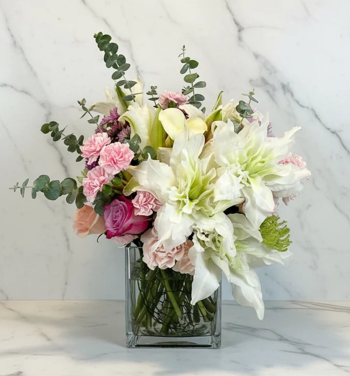 Elegant floral arrangement with white lilies, pink roses, and green foliage in a clear glass vase on marble surface