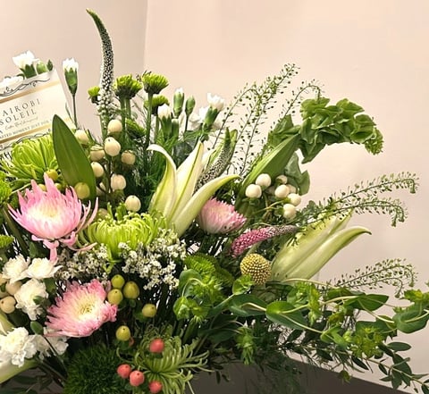 Vibrant floral arrangement with pink protea, white lilies, babys breath, and mixed green foliage in a dark vase