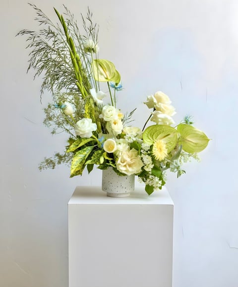 Elegant white and green floral arrangement in a speckled vase on a white pedestal against a white wall