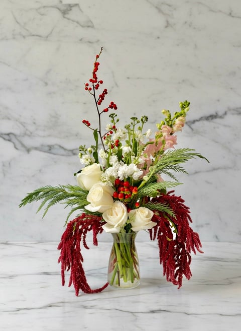 Elegant floral arrangement with white roses, red berries, burgundy amaranthus, and green foliage in a glass vase on marble background