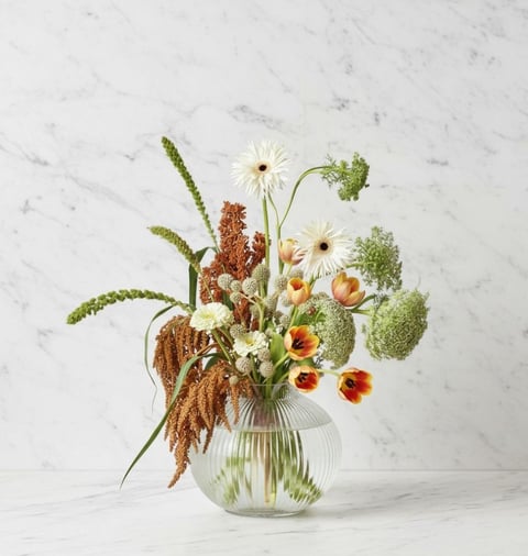 Mixed flower arrangement in glass vase featuring daisies, tulips, dried grains, and green foliage on marble background