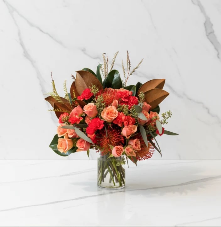 Coral and red floral arrangement in a clear glass vase with roses, carnations, and greenery on a marble background
