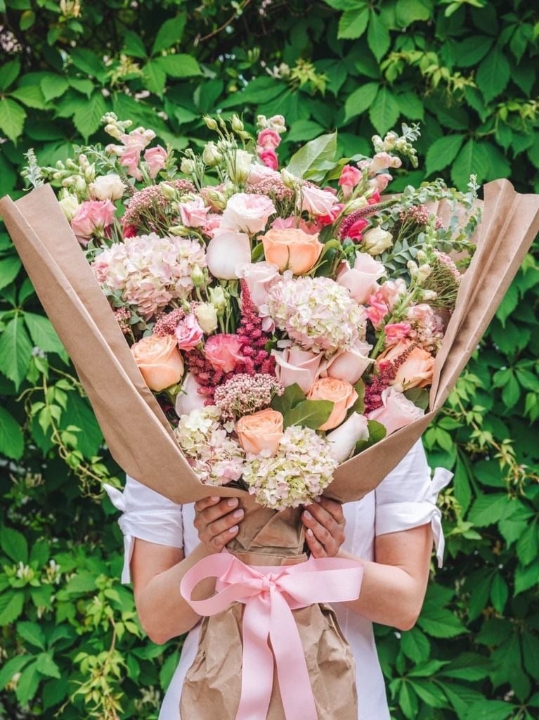 Person holding large bouquet of pink and white flowers wrapped in kraft paper with green ivy backdrop