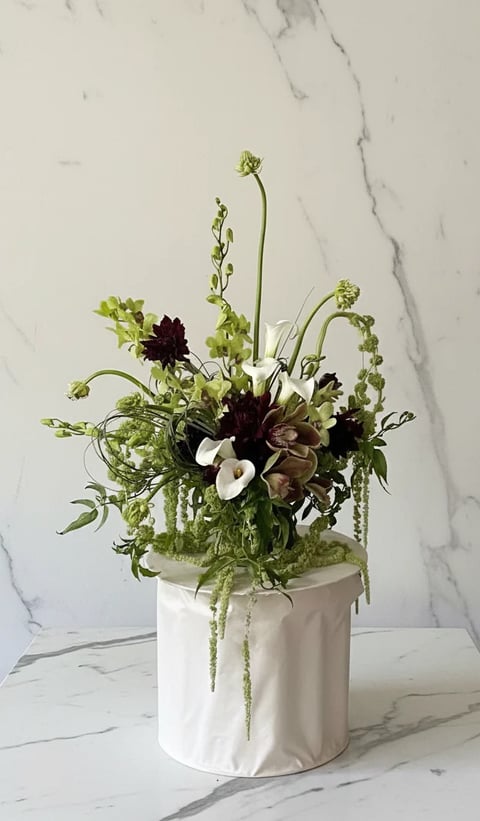 Floral arrangement in white pot with dark burgundy flowers, white blooms, cascading greenery, and curving stems on marble surface