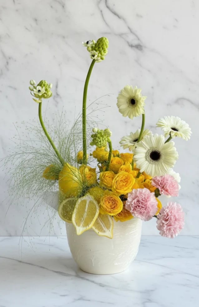 Floral arrangement in white ceramic vase with yellow roses, white gerbera daisies, pink carnations, and green accent stems on marble background