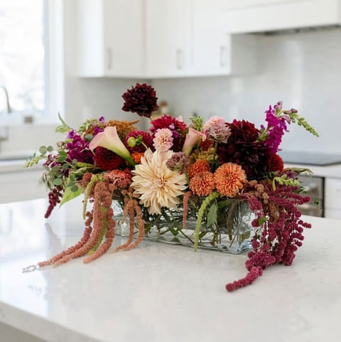Lush floral arrangement with burgundy dahlias, coral mums, and trailing amaranthus in a glass vase on a kitchen counter