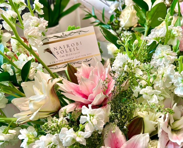 A Nairobi Love Letter perfume box nestled among white babys breath flowers and pink roses in a floral arrangement