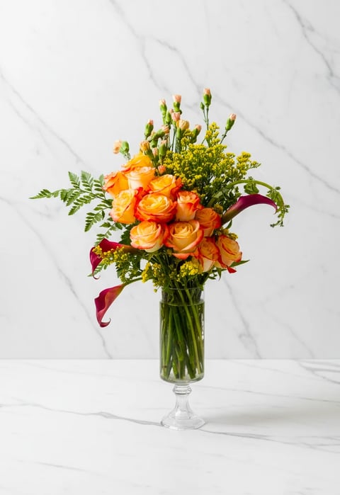A vibrant floral arrangement in a clear glass vase featuring orange roses, burgundy calla lilies, green lisianthus, and mixed greenery on a marble background
