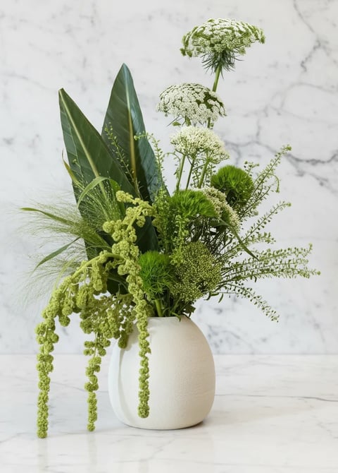 Green floral arrangement with lush foliage, delicate fern fronds, and white flowers in a cream ceramic vase on marble surface